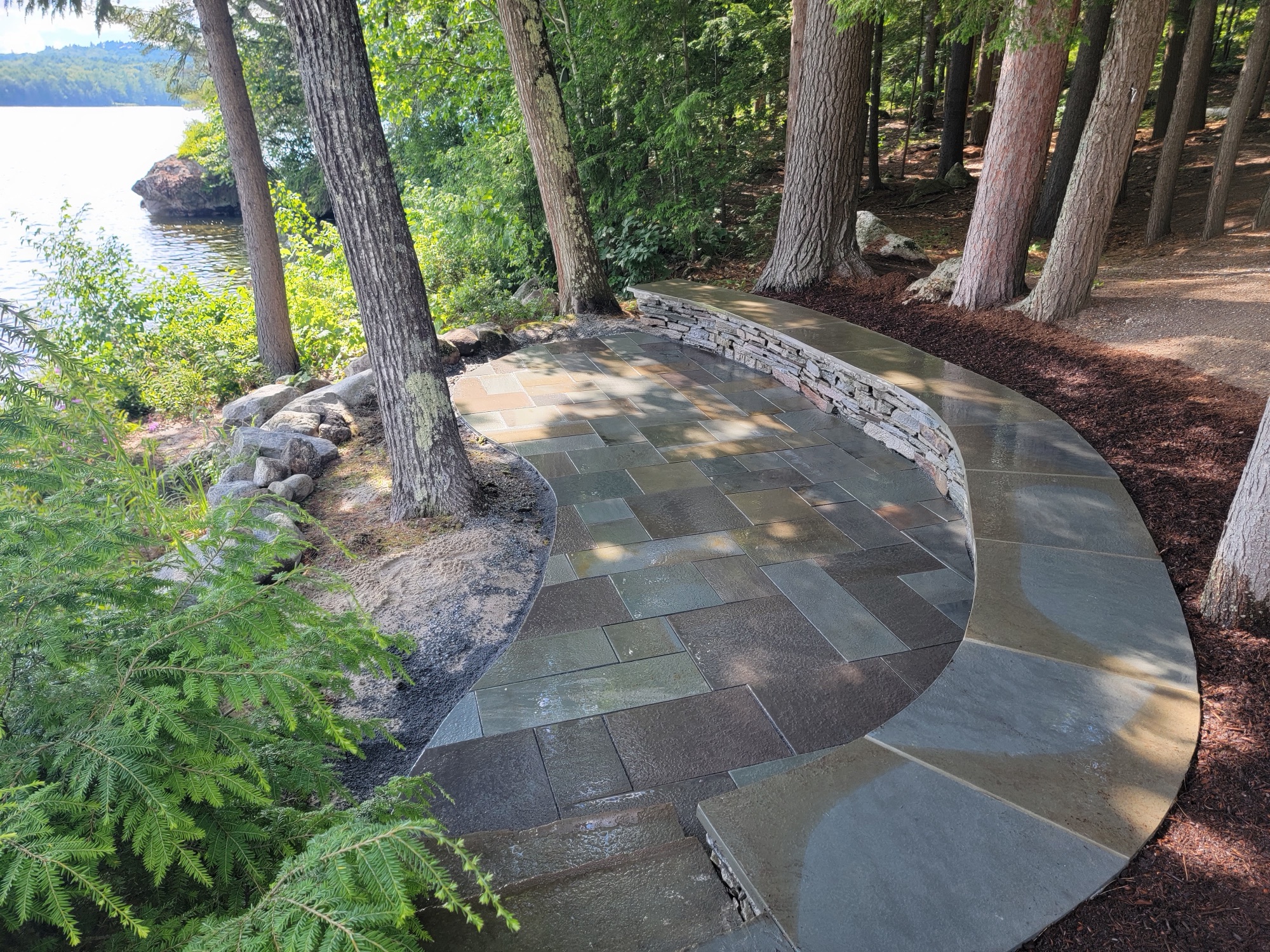 Curved bluestone patio with low stacked-stone retaining wall, lake glimpsed through pines