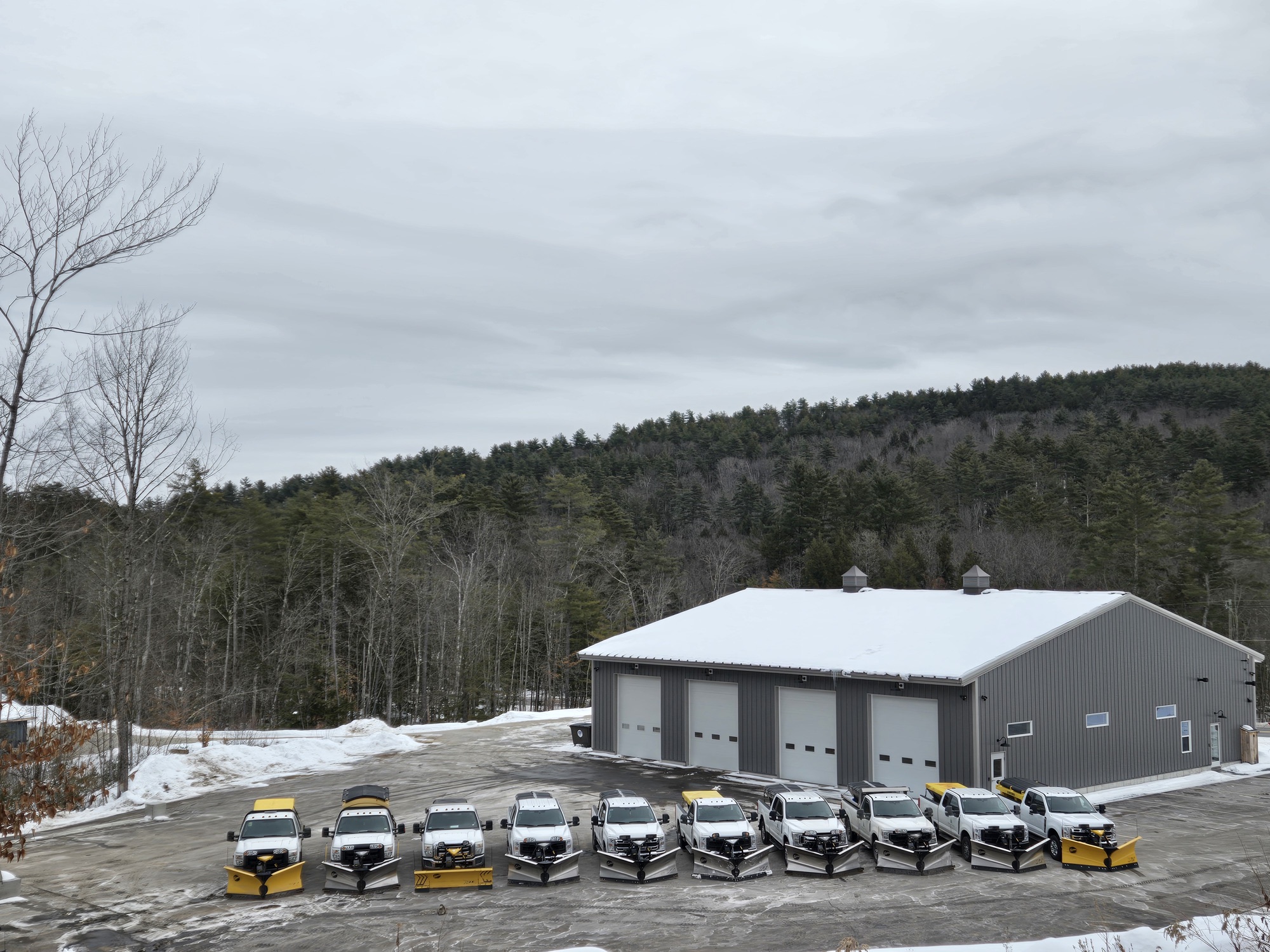 Fleet of plow trucks lined up at the company shop with snowy hills behind