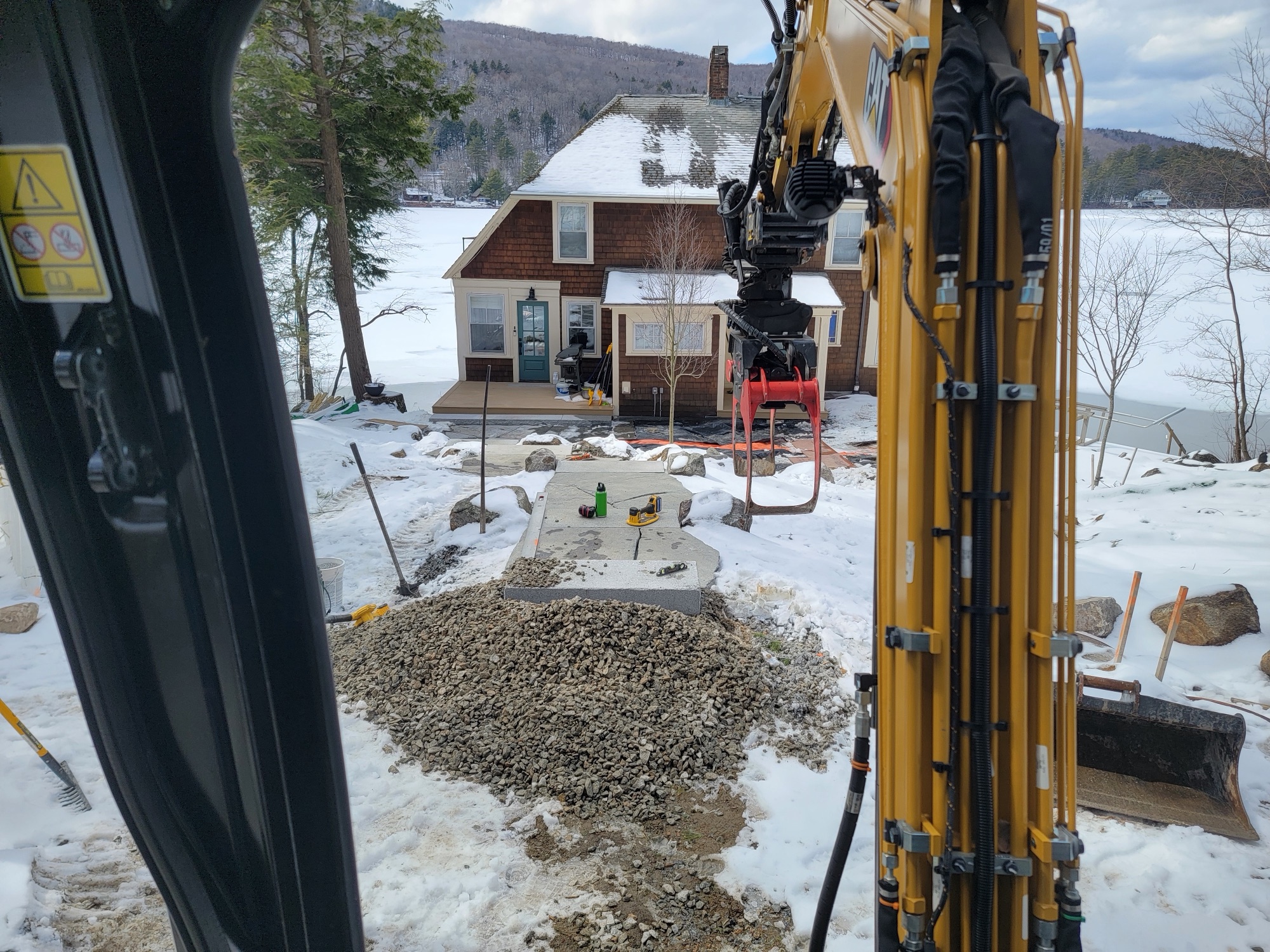 Winter shot from inside an excavator cab toward a snow-covered lakefront cottage