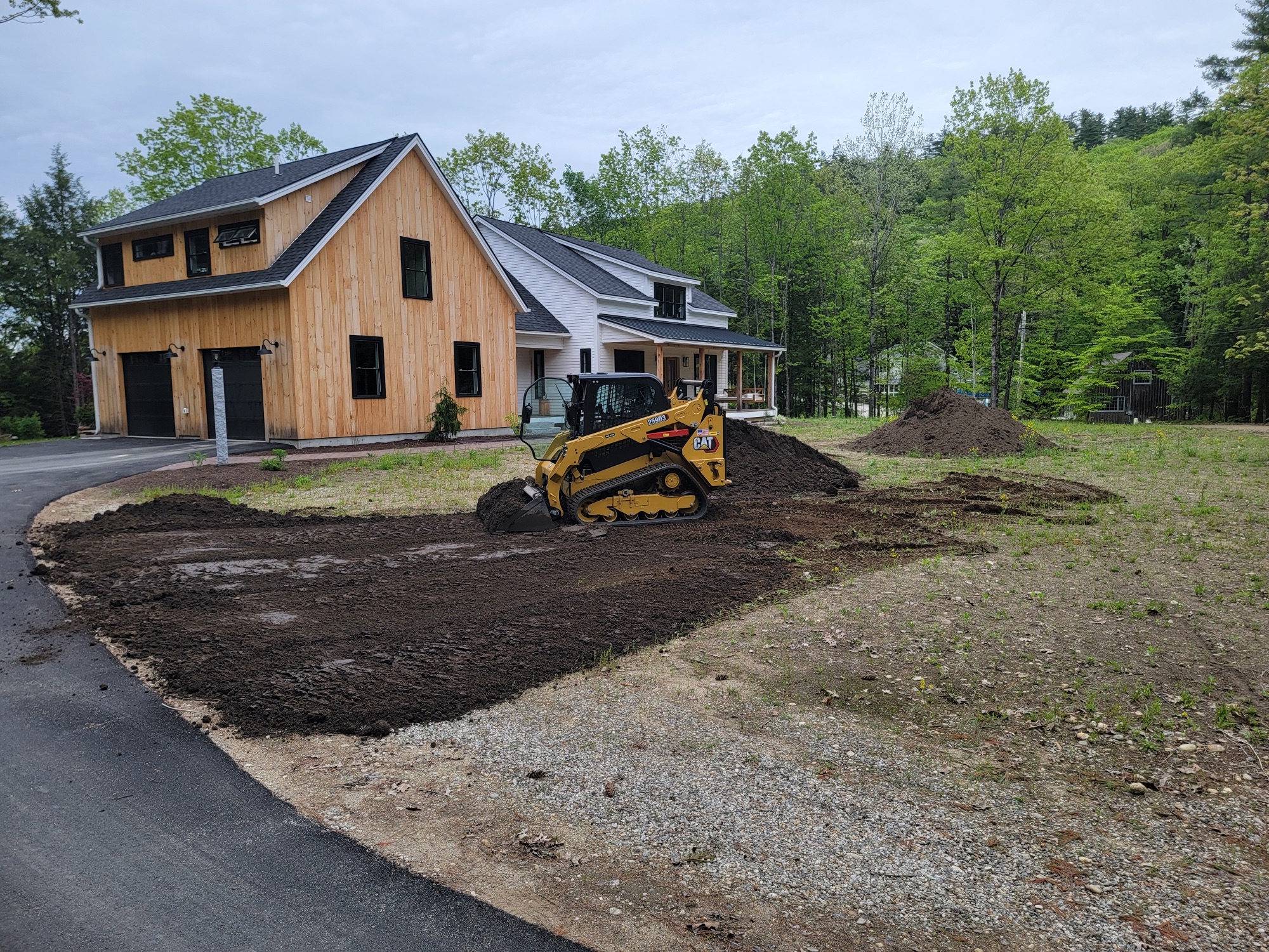 Compact track loader spreading topsoil along the front of a barn-style home